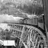 White Pass and Yukon train on a trestle, with mining operation below the bridge. 1906.